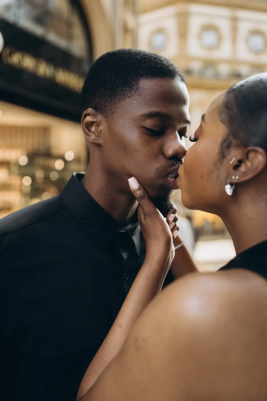 A close-up kiss, her fingers lifting his chin and a drop earring catching the light, with Galleria shopfronts blurred behind.