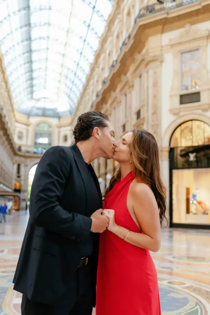 A couple kisses while holding hands at chest height, the long glass roof of the Galleria stretching away behind them.