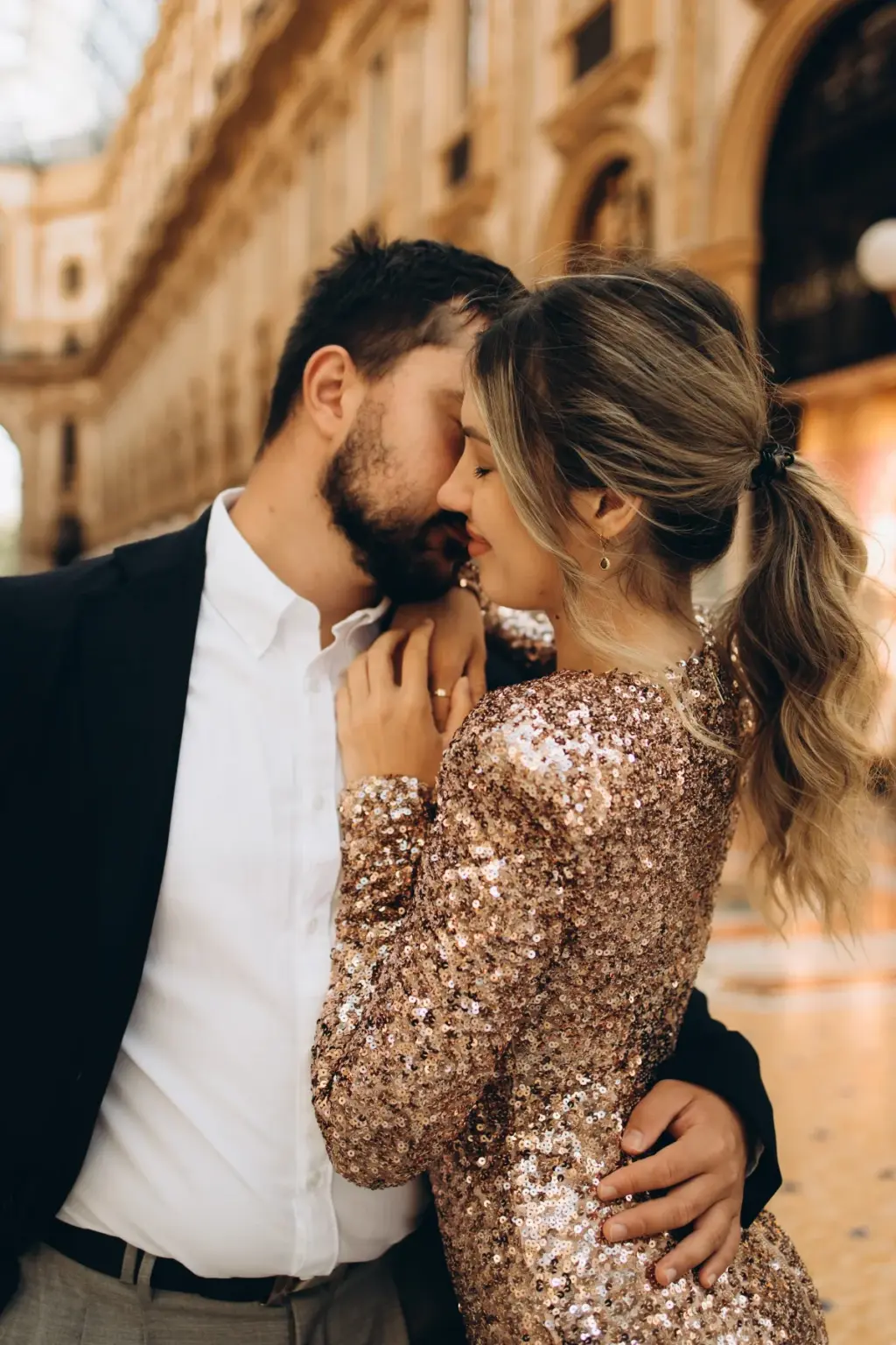 A woman with a ponytail and glittering sleeves leans into her partner for a near kiss among the arcades of the Galleria.