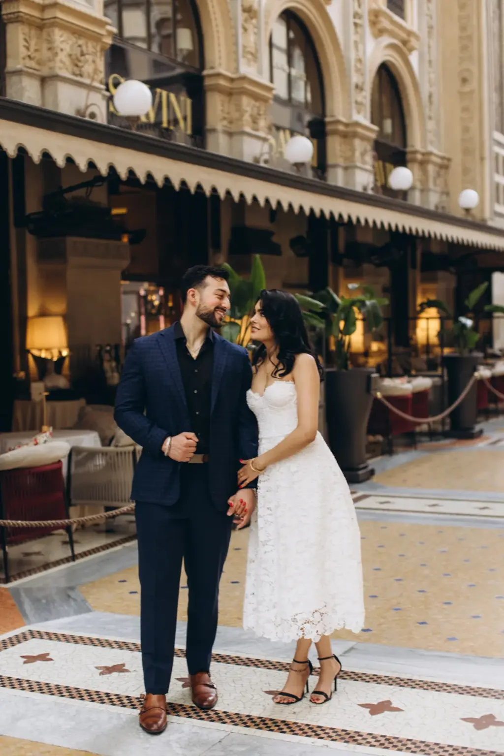 Holding hands on the mosaic floor, they laugh together in front of a café terrace with globe lamps inside the Galleria.