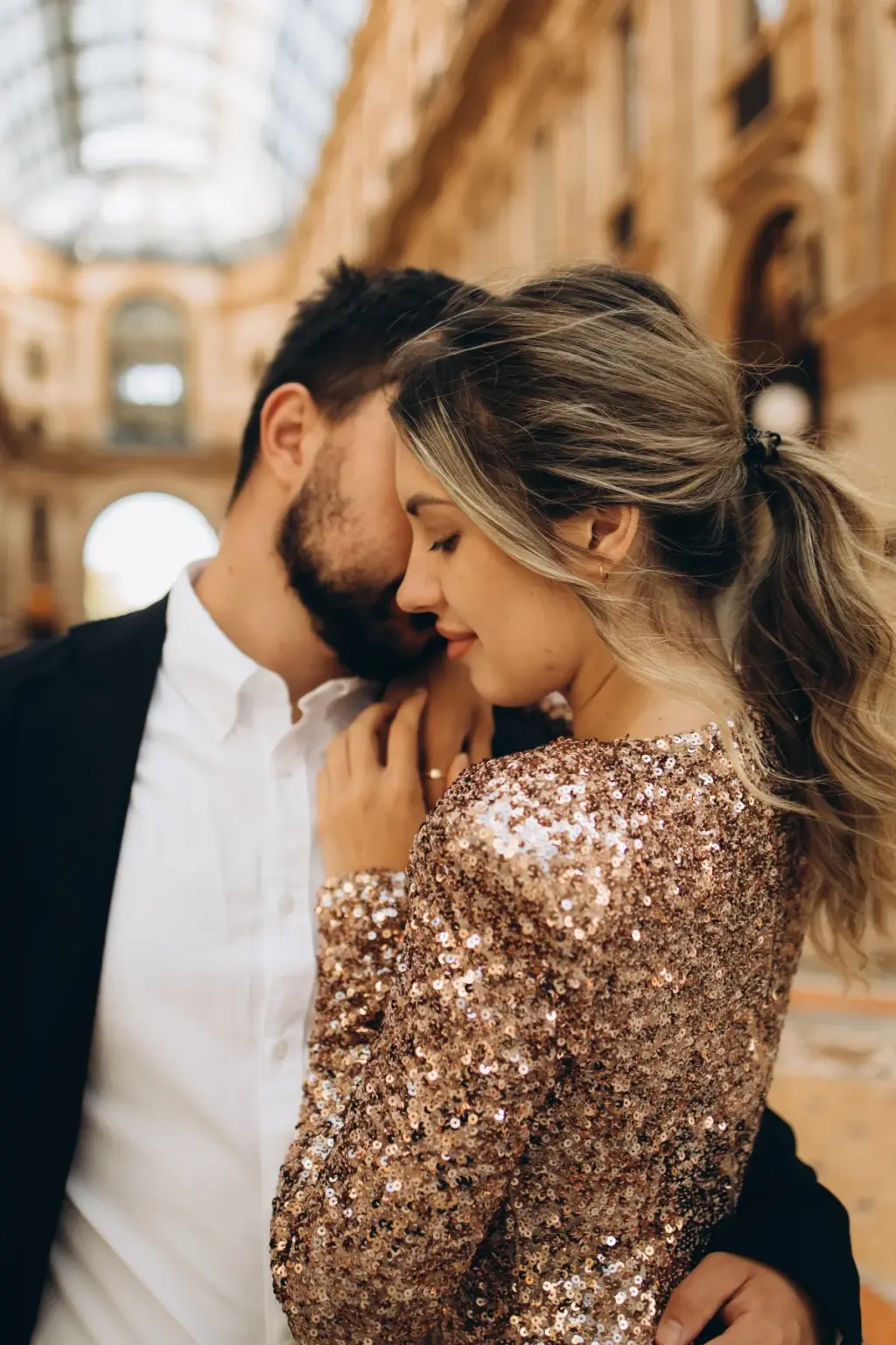 Close-up of a bearded man pressing his face to his partner's cheek as she closes her eyes, the Galleria glass roof glowing behind.
