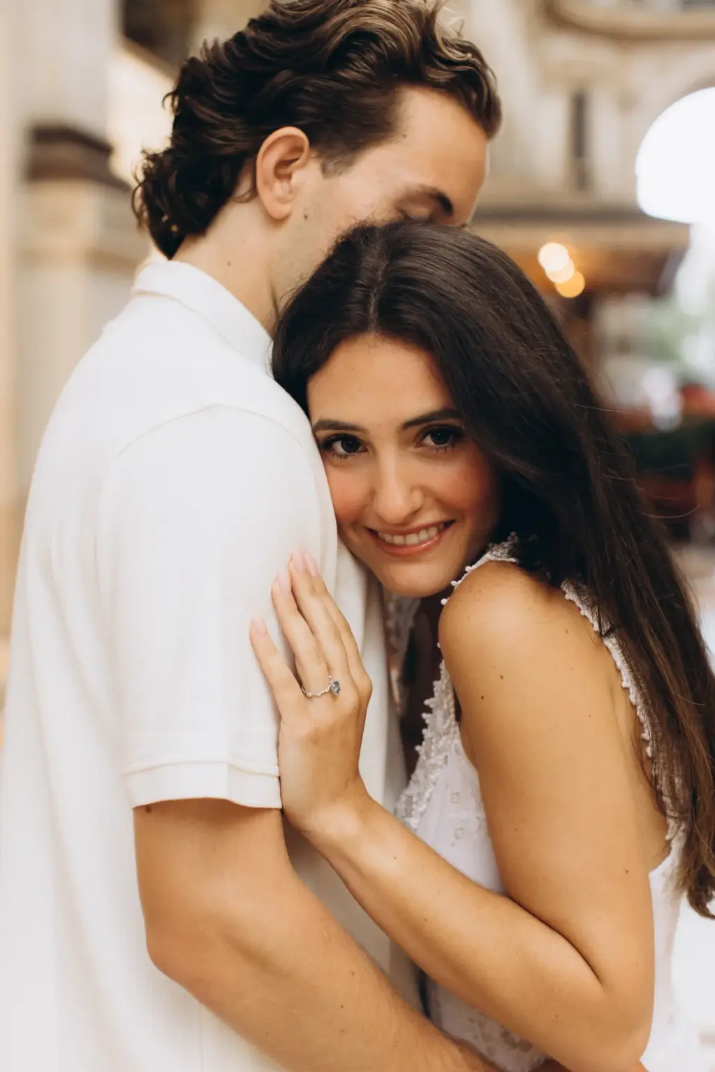 A woman with dark hair smiles at the camera as she hugs her partner, who presses his face into her hair inside the Galleria.