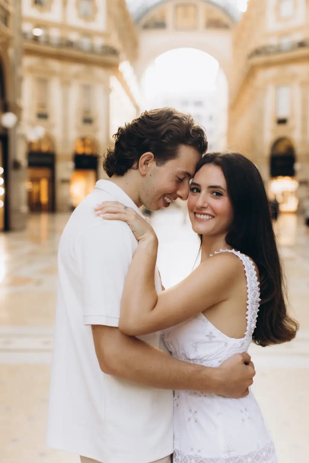 Under the glass arch of the Galleria Vittorio Emanuele II, a man presses his forehead to his smiling partner's temple.
