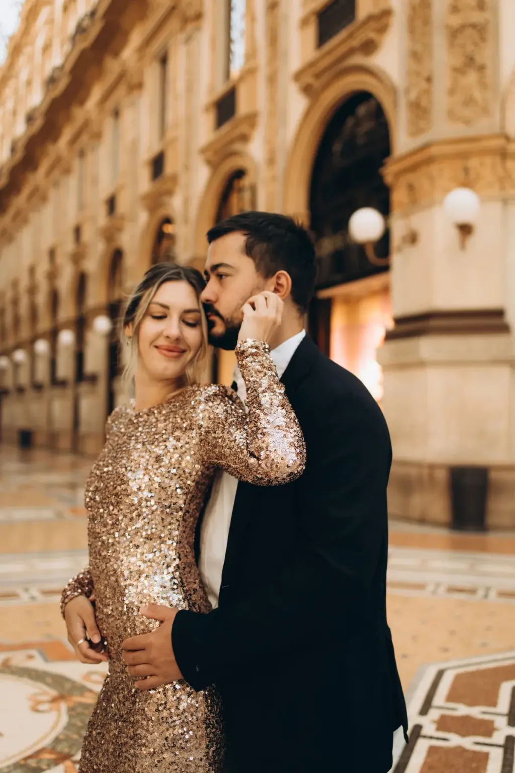 Smiling, a woman in a sparkling dress holds her partner's hand to her cheek as he kisses her hair in the Galleria Vittorio Emanuele II.