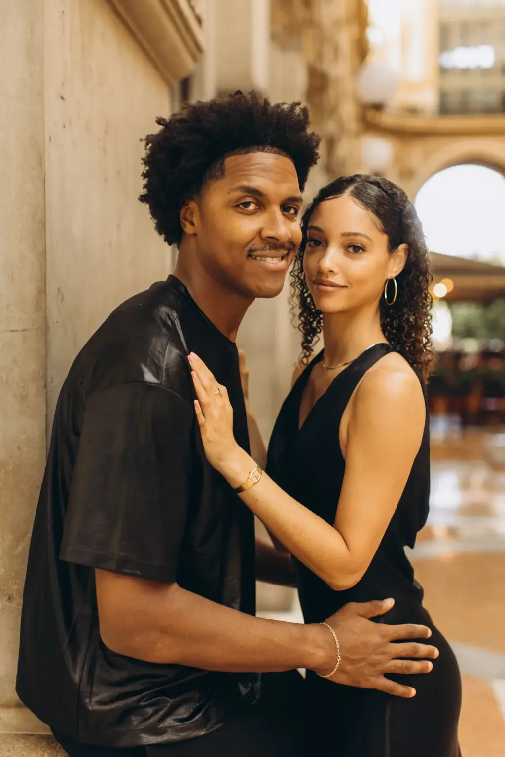 A man and woman in black lean against a pale wall in the Galleria, her hand flat on his chest as they both smile at the camera.