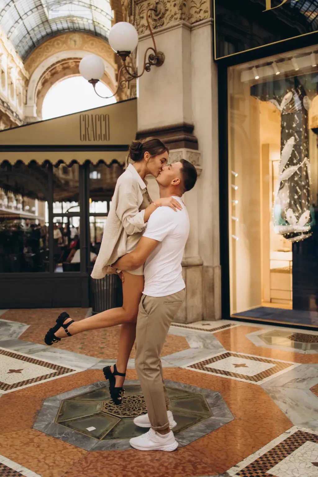 In the Galleria Vittorio Emanuele II, a woman in a beige blazer kicks one heel up as her partner lifts her into a kiss on the mosaic floor.