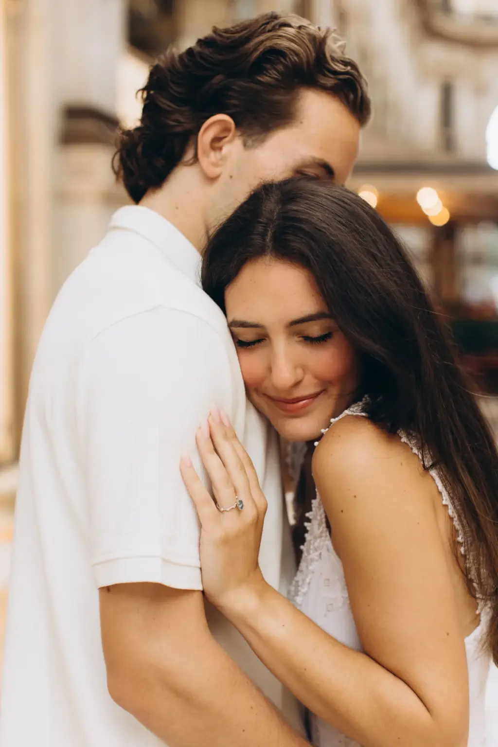 Eyes closed, a woman rests her head on her partner's chest, a ring on the hand that holds his shoulder, inside the Galleria.