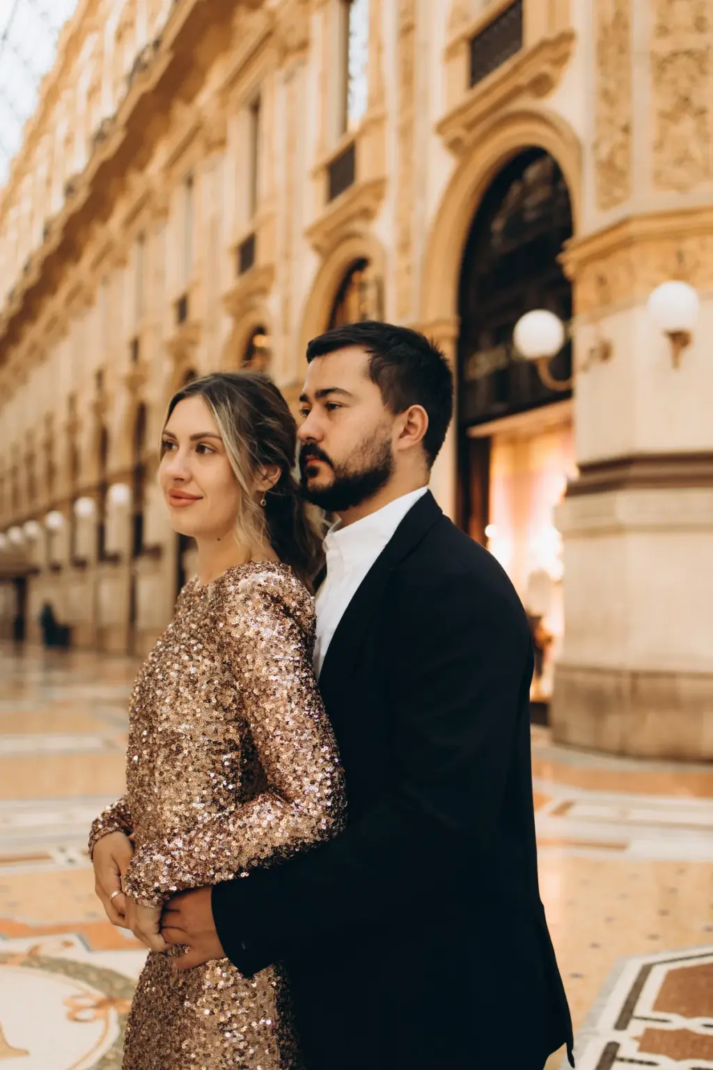 A woman in a rose-gold sequin dress stands with her partner behind her, his face turned to the side, inside the Galleria.