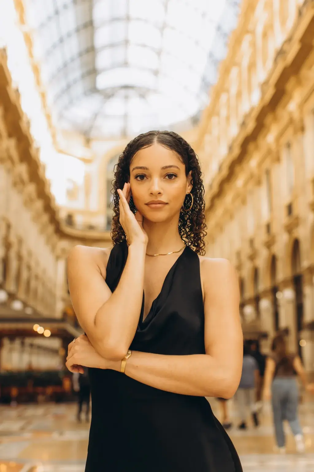A woman in a black halter dress poses alone in the Galleria, one hand at her cheek and her arm folded under the glass roof.