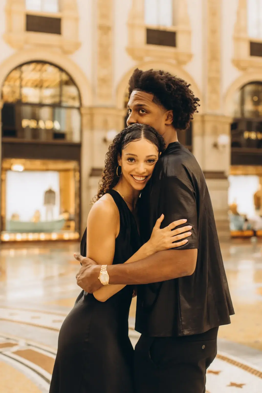 A smiling woman is wrapped in her partner's arms on the mosaic floor of the Galleria, a gilded shopfront glowing behind them.