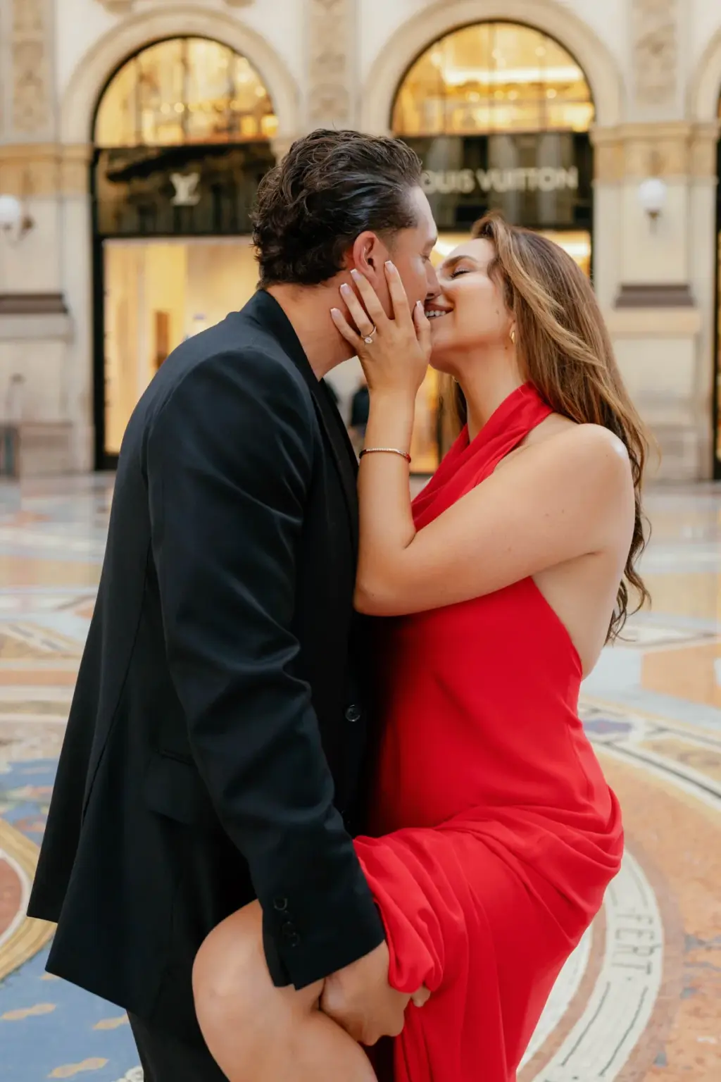 Laughing, she cups his face and hooks a leg around him on the Galleria's mosaic floor, red dress swirling around her knees.