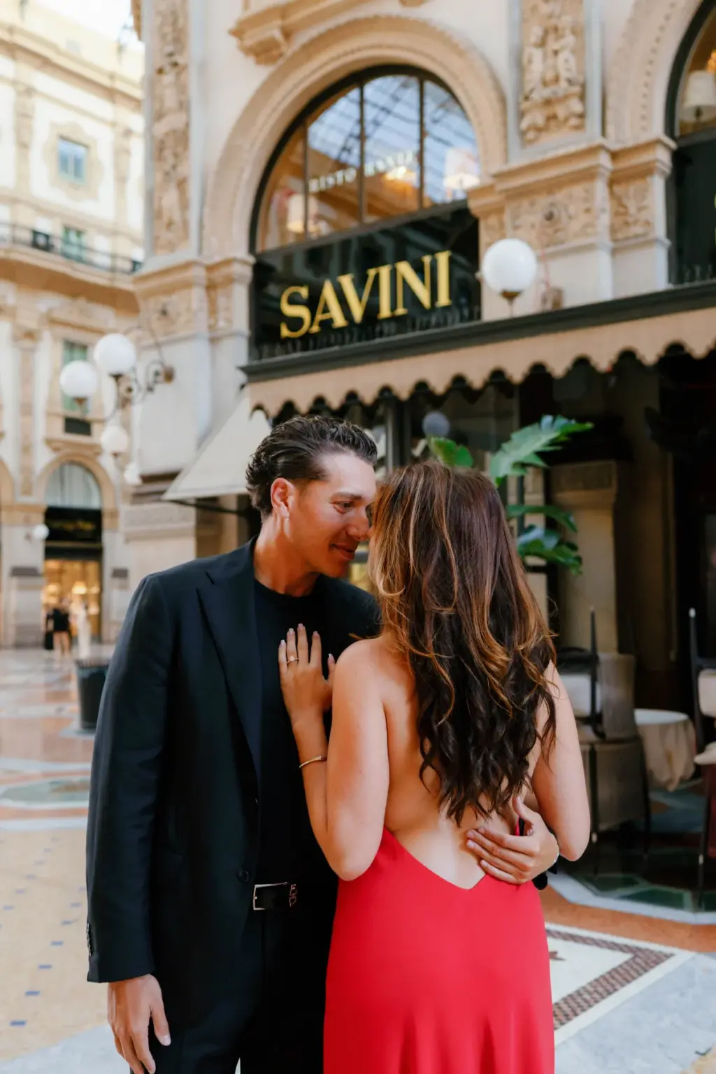 A woman in a backless red dress leans into her partner in front of the Savini restaurant sign inside the Galleria in Milan.