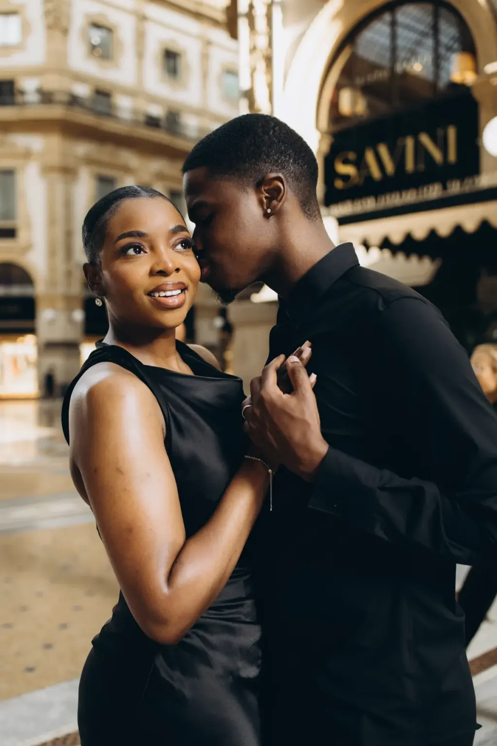 He kisses her temple as she smiles off to the side, the gold Savini lettering glowing above the Galleria café behind them.