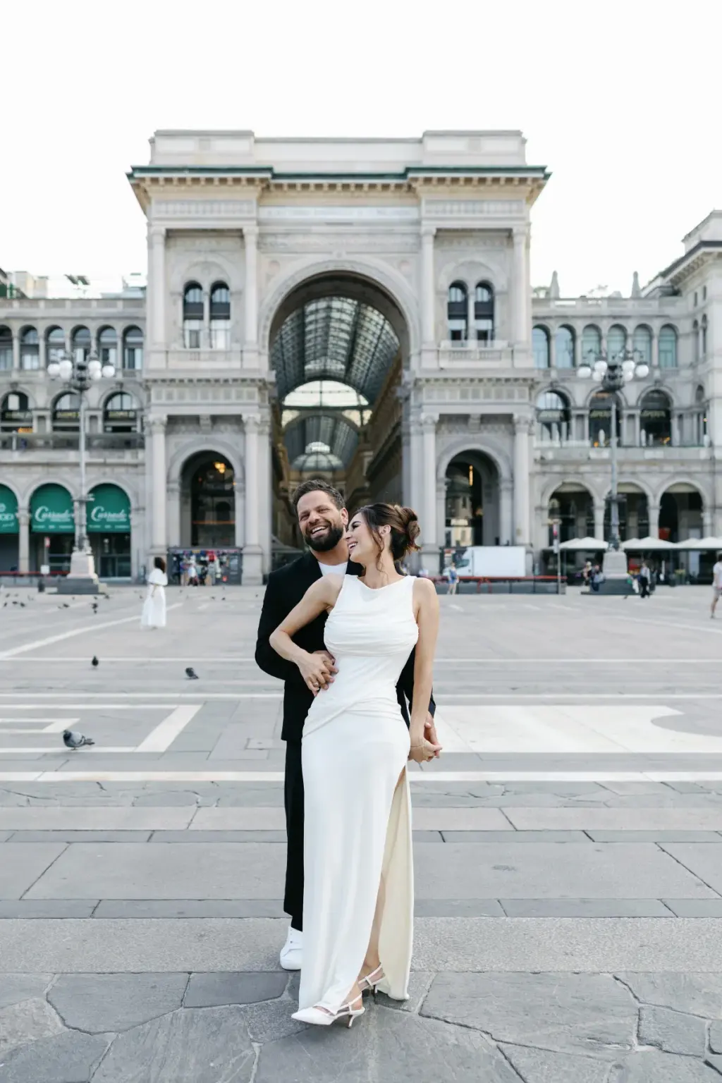 He hugs her from behind on the Duomo square, with the triumphal arch entrance of Galleria Vittorio Emanuele II behind them.
