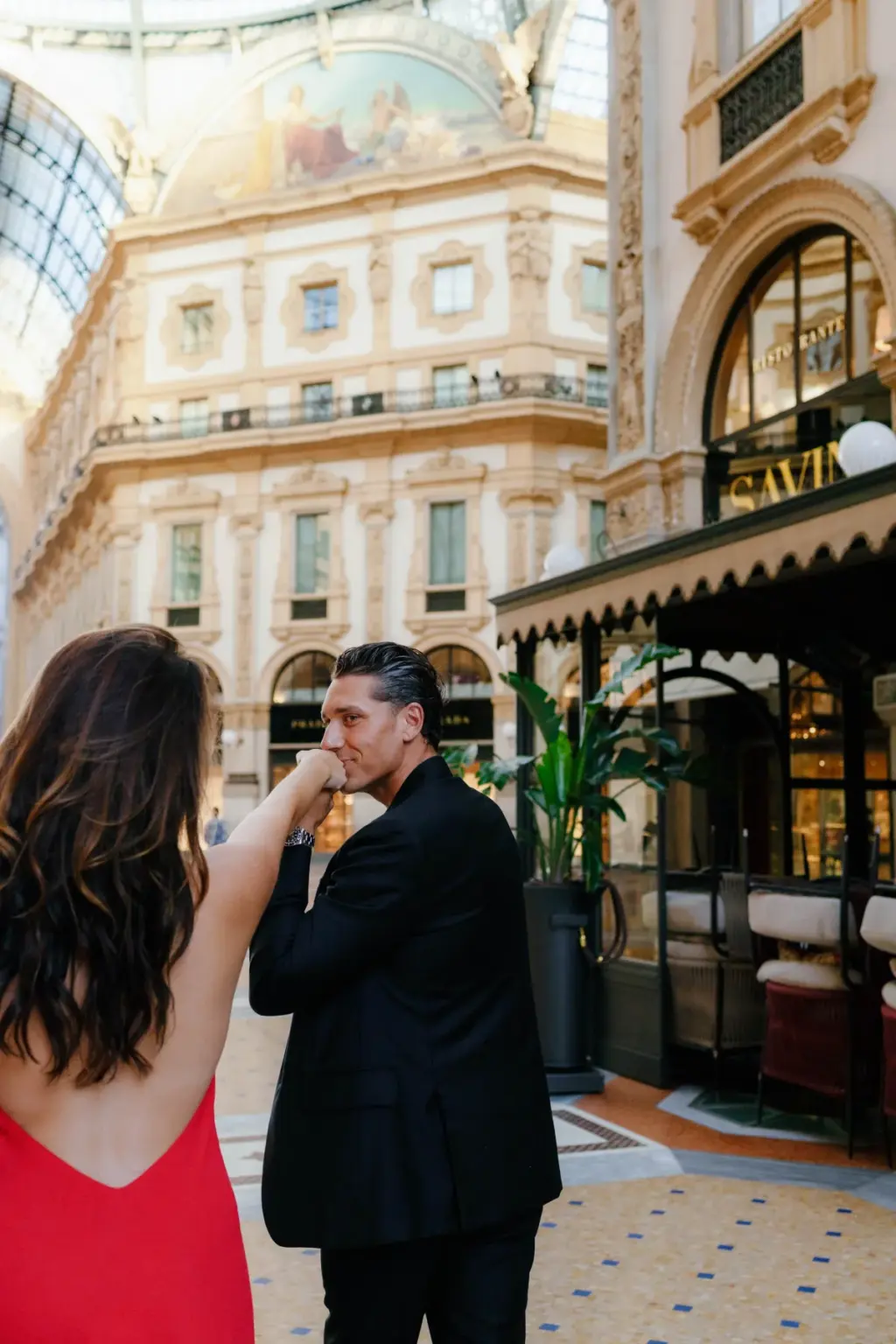 He kisses her hand as she leads him past the Savini café terrace, the frescoed Galleria dome glowing overhead.