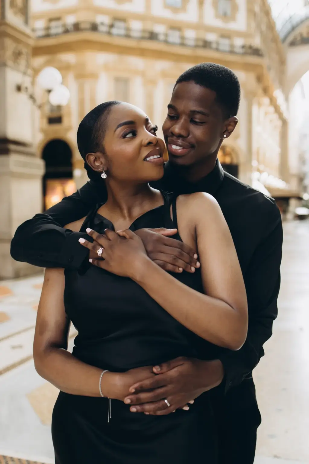 He wraps his arms around her from behind and she looks up at him, her hand with an engagement ring over his, in the Galleria.