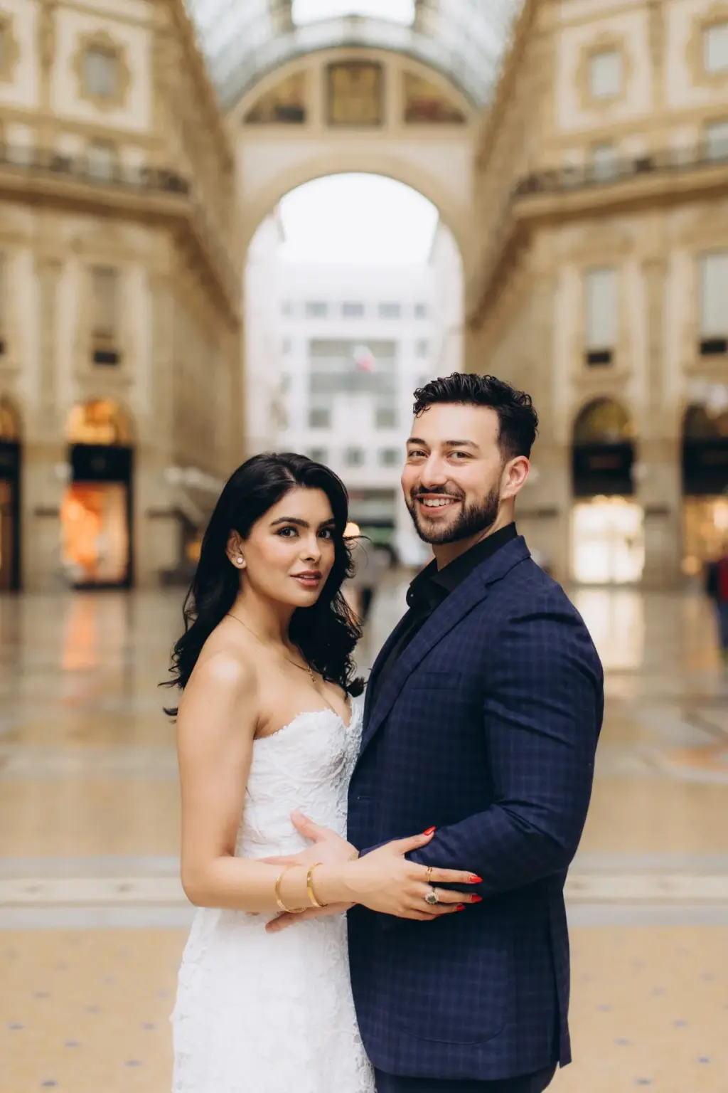 Standing close in the Galleria Vittorio Emanuele II, the couple smiles at the camera with its tall arched exit glowing behind.