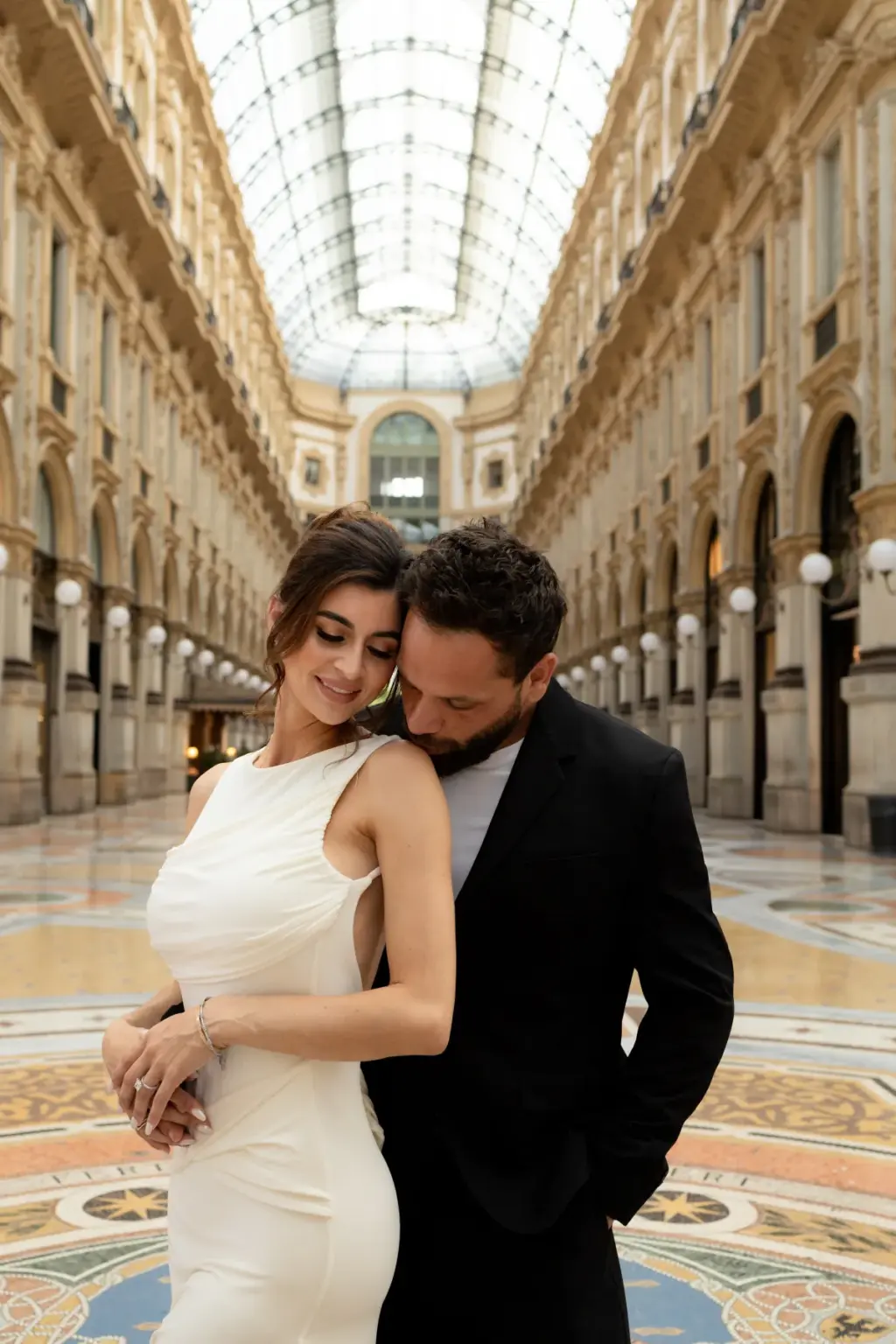 Under the glass vault of Galleria Vittorio Emanuele II, he kisses her shoulder as she smiles down, mosaic floor at their feet.