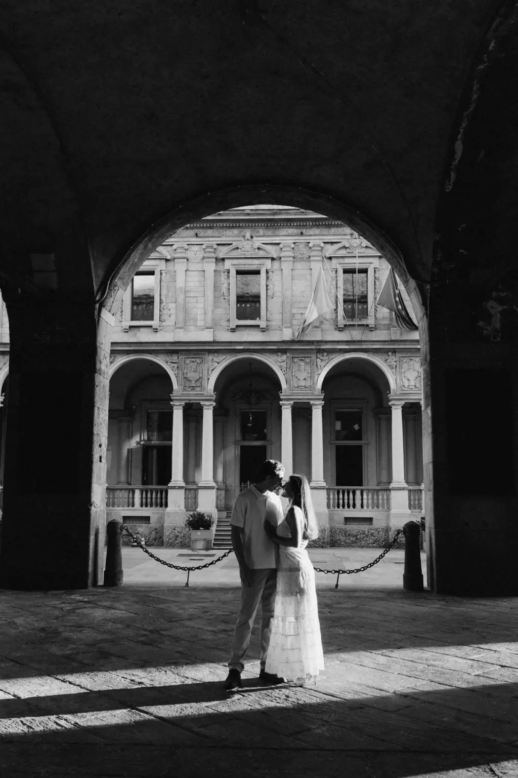 In black and white, a couple kisses under a deep stone arch that frames the colonnaded courtyard of the Loggia dei Mercanti.