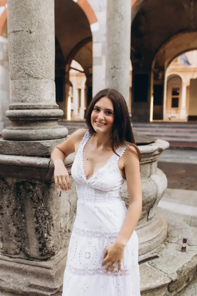 A smiling woman in a white eyelet dress leans on the carved stone well in the Loggia dei Mercanti courtyard, columns behind her.