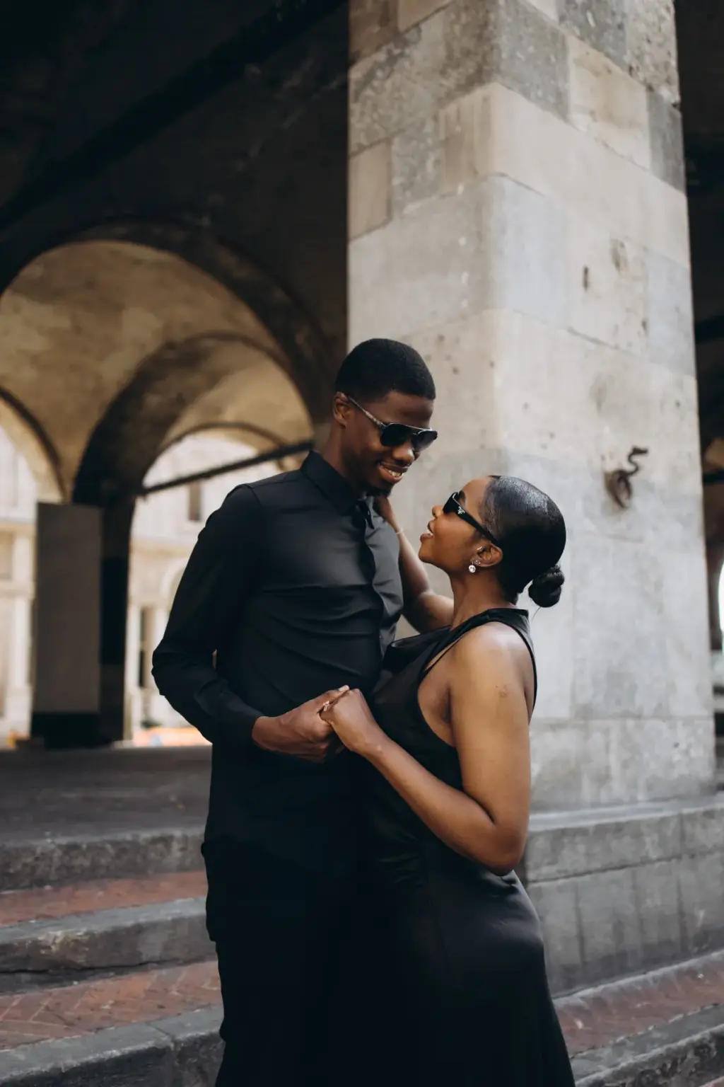 Holding both hands, the couple shares a smile in their sunglasses by the arcade's worn steps and heavy stone pillar.