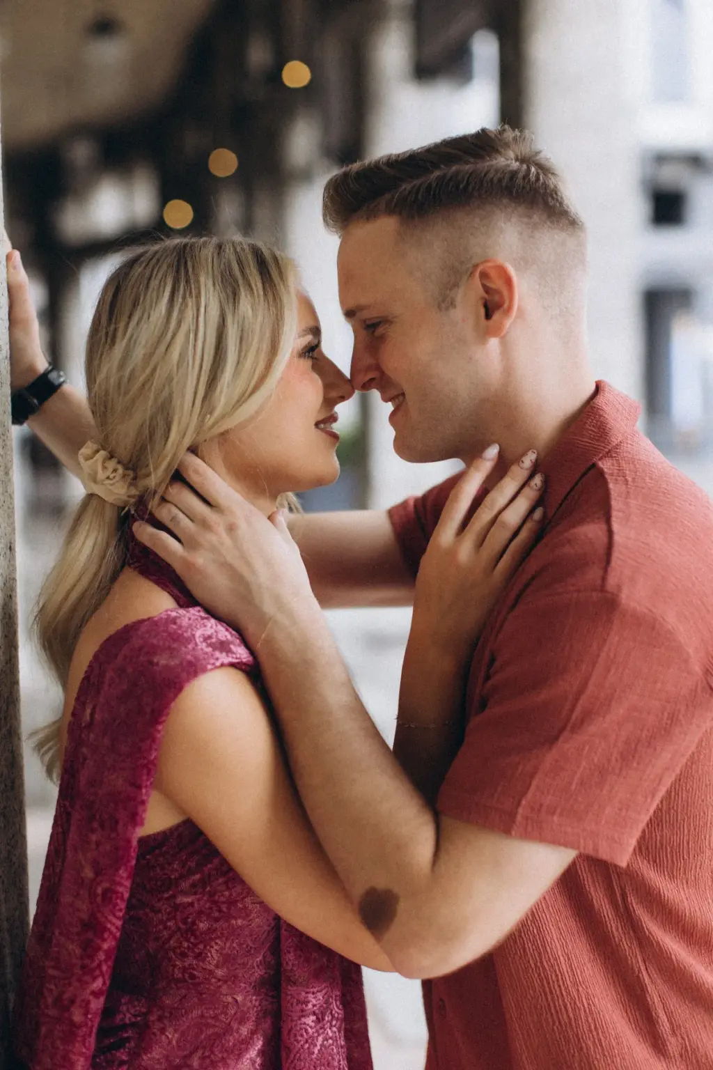 Under a stone arcade with soft lights glowing behind, she holds his neck in both hands as they stand nose to nose.