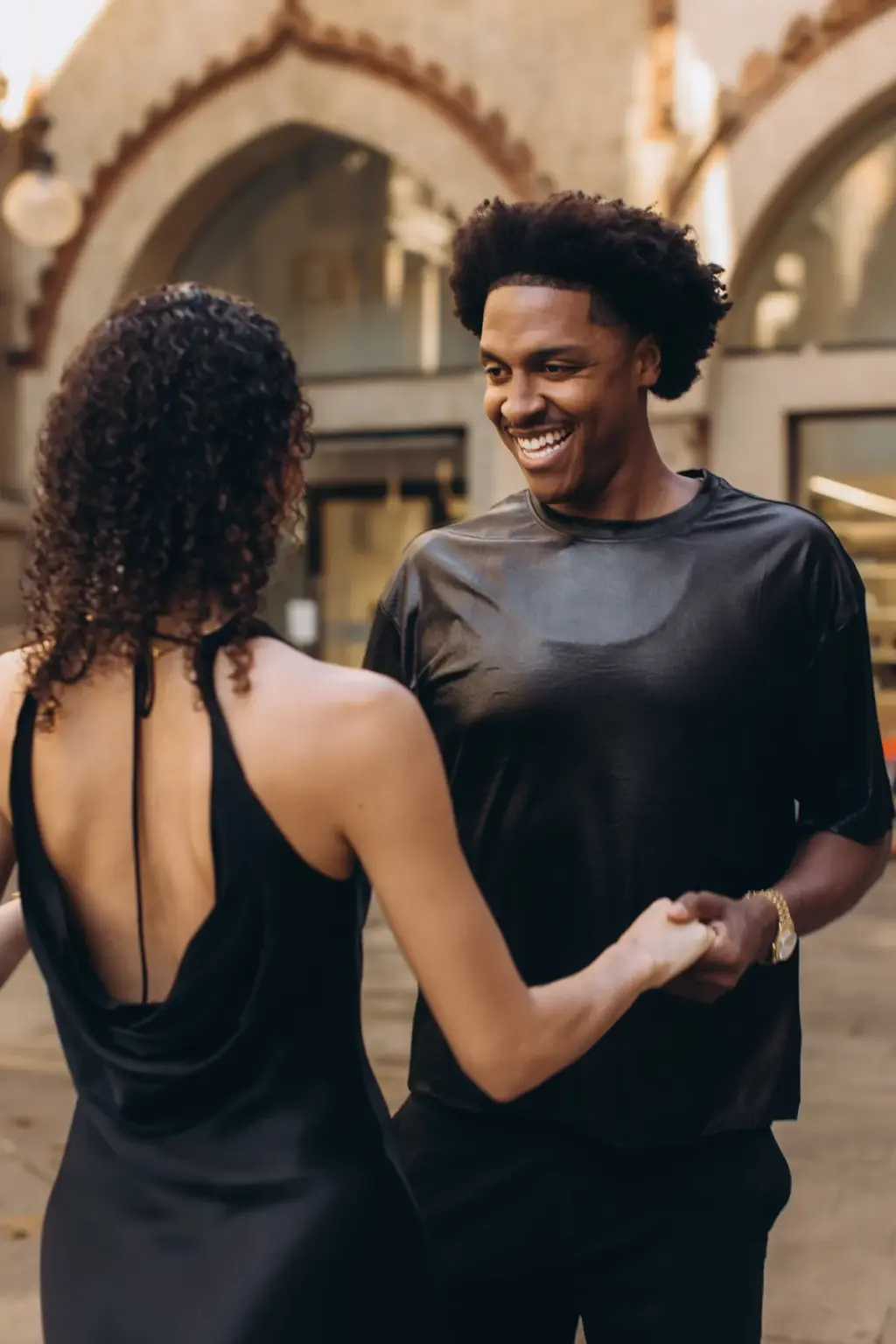 A smiling man holds his partner's hand as she twirls away from him under brick-trimmed stone arches on a Milan street.