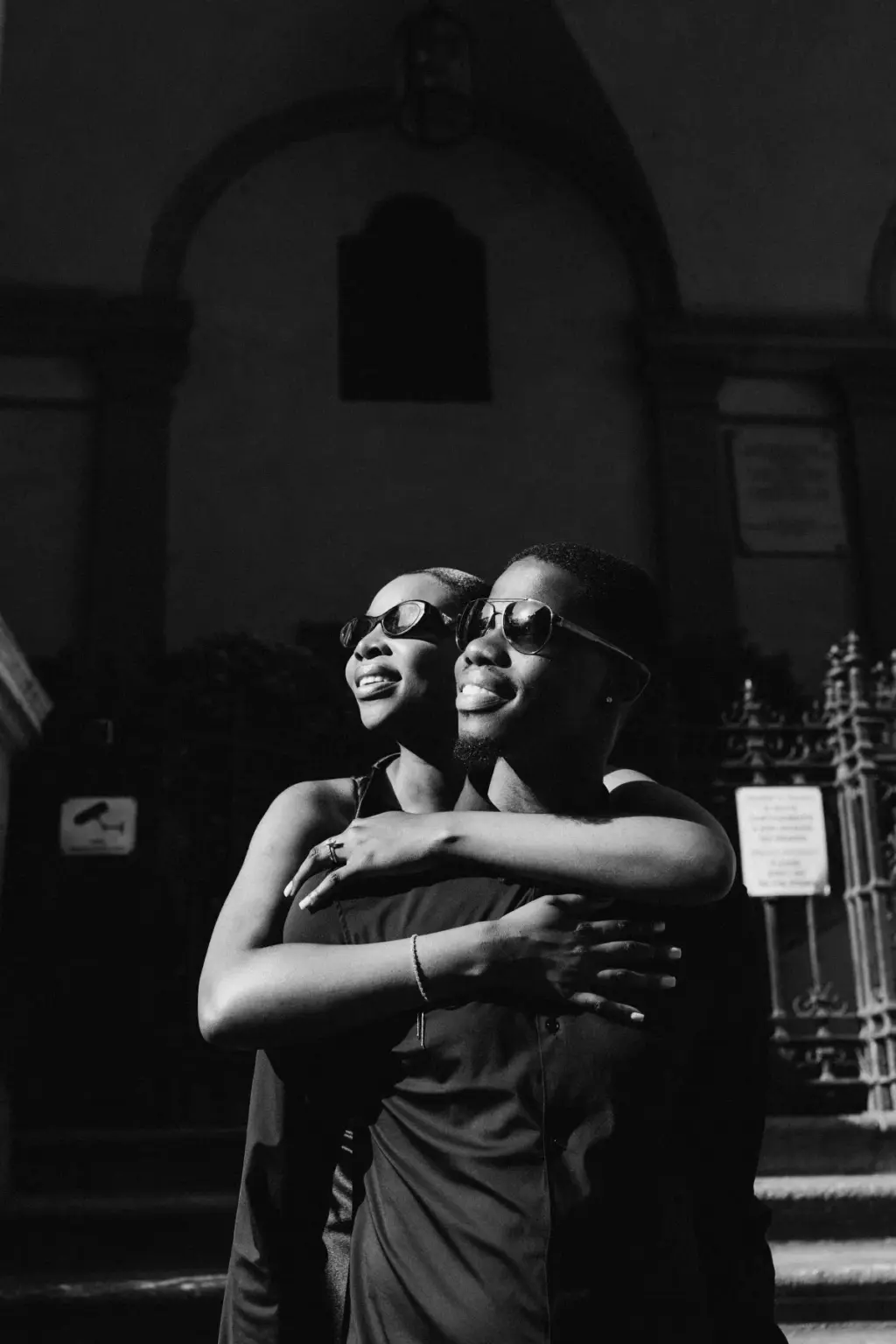 Black-and-white frame of the couple in sunglasses, her arms around him from behind as they turn their faces to the sun.