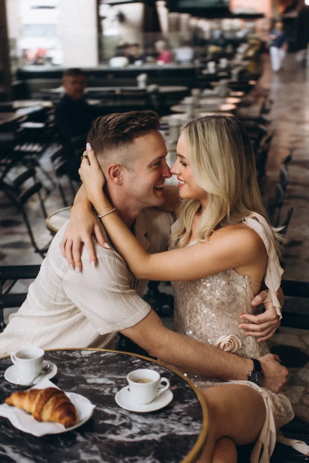 At a marble café table with two espressos and a croissant, the couple holds each other close, nose to nose and smiling.