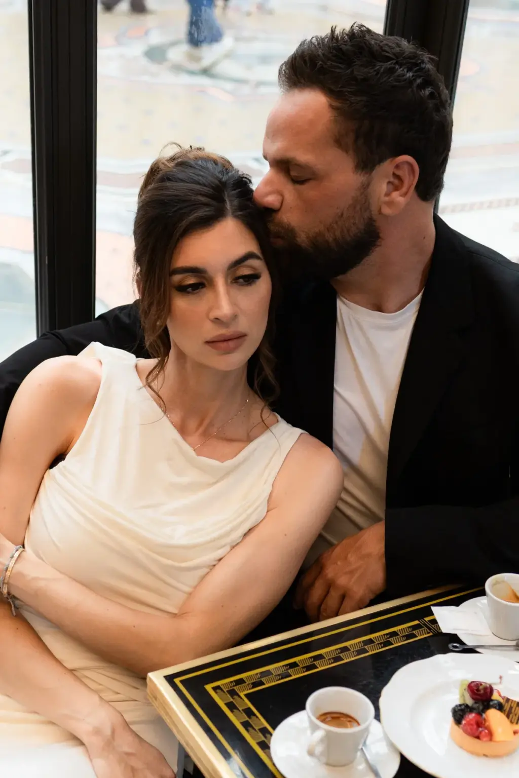 At a café table with espresso cups and small pastries, he kisses the top of her head as she gazes off past the window.