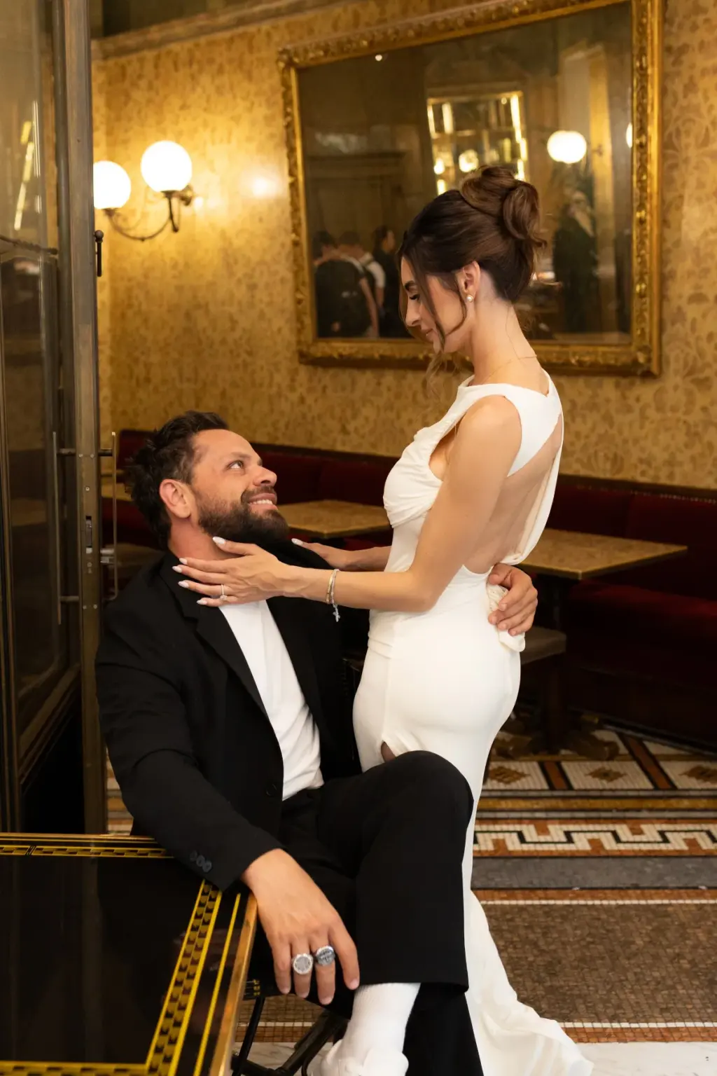 Inside a café with a gilded mirror and red banquettes, he sits laughing up at her as she stands close in her white dress.