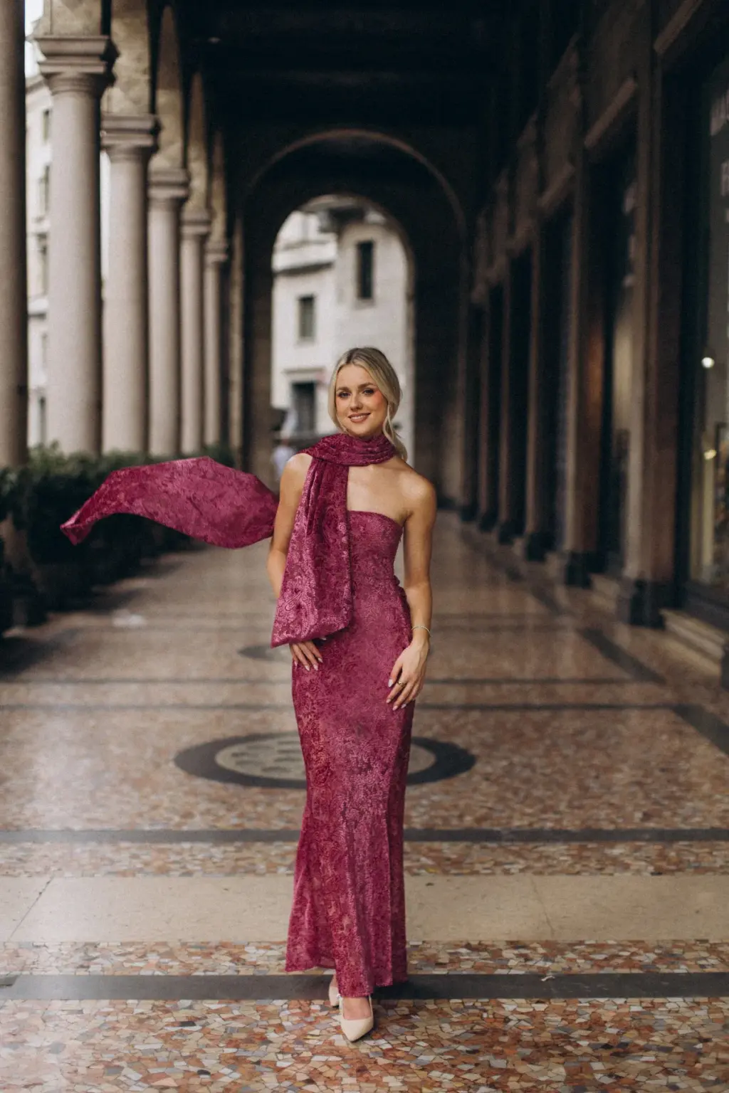 Her raspberry scarf flies out to the side as she stands alone in a long marble-floored colonnade in central Milan.