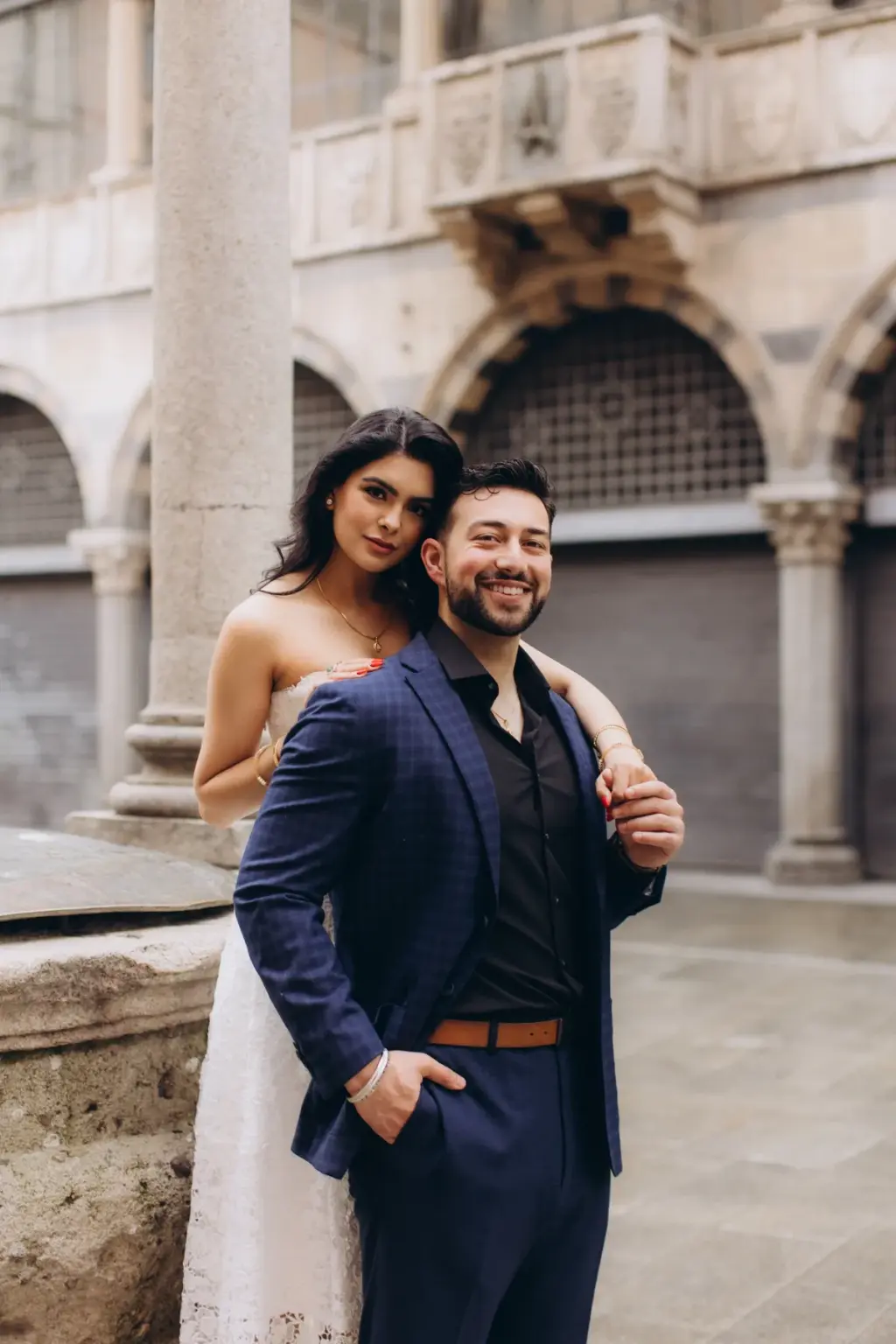 Draping an arm over his shoulder from behind, she poses with him beside an old stone well head in a colonnaded Milan loggia.