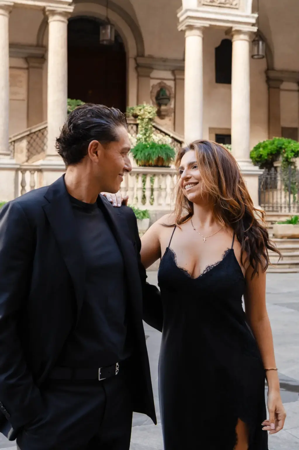 Now in a black lace slip dress, she smiles at him in a colonnaded palazzo courtyard with potted plants on the balustrade.