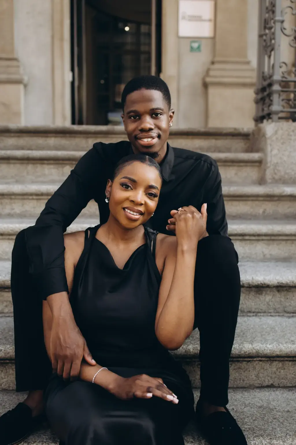 Sitting together on the stone steps of a Milan palazzo, he hugs her from the step above as both smile at the camera.