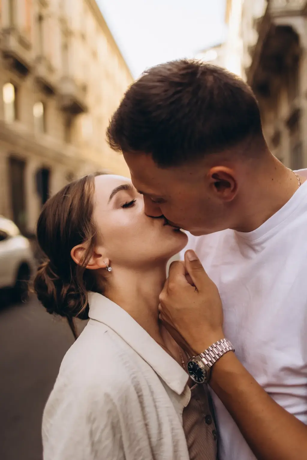 Eyes closed, a woman in a cream jacket is kissed on the cheek by her partner, his hand at her neck, on a Milan side street.