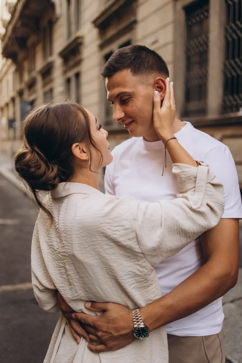 A woman in a linen jacket cups her partner's face as he smiles down at her, holding her waist on a quiet Milan side street.