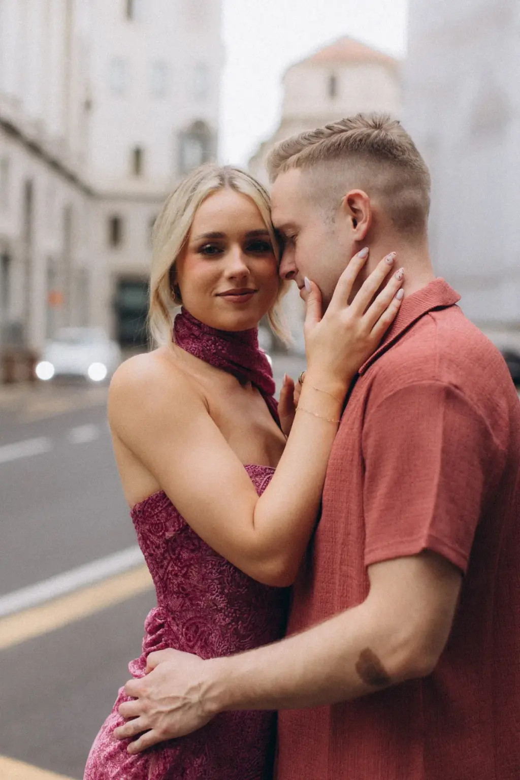In a raspberry lace dress with a matching neck scarf, she cups his face as he leans into her on a Milan side street.