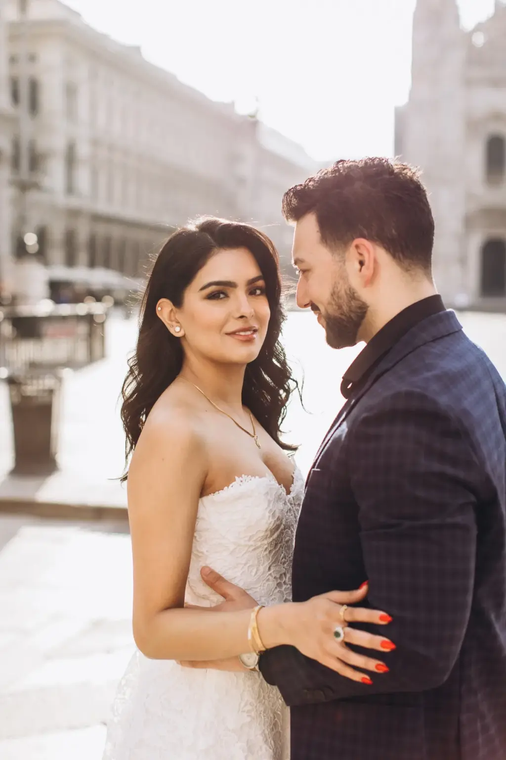 She glances over at the camera with a soft smile while he looks at her, his arm around her waist on a sunlit Milan square.