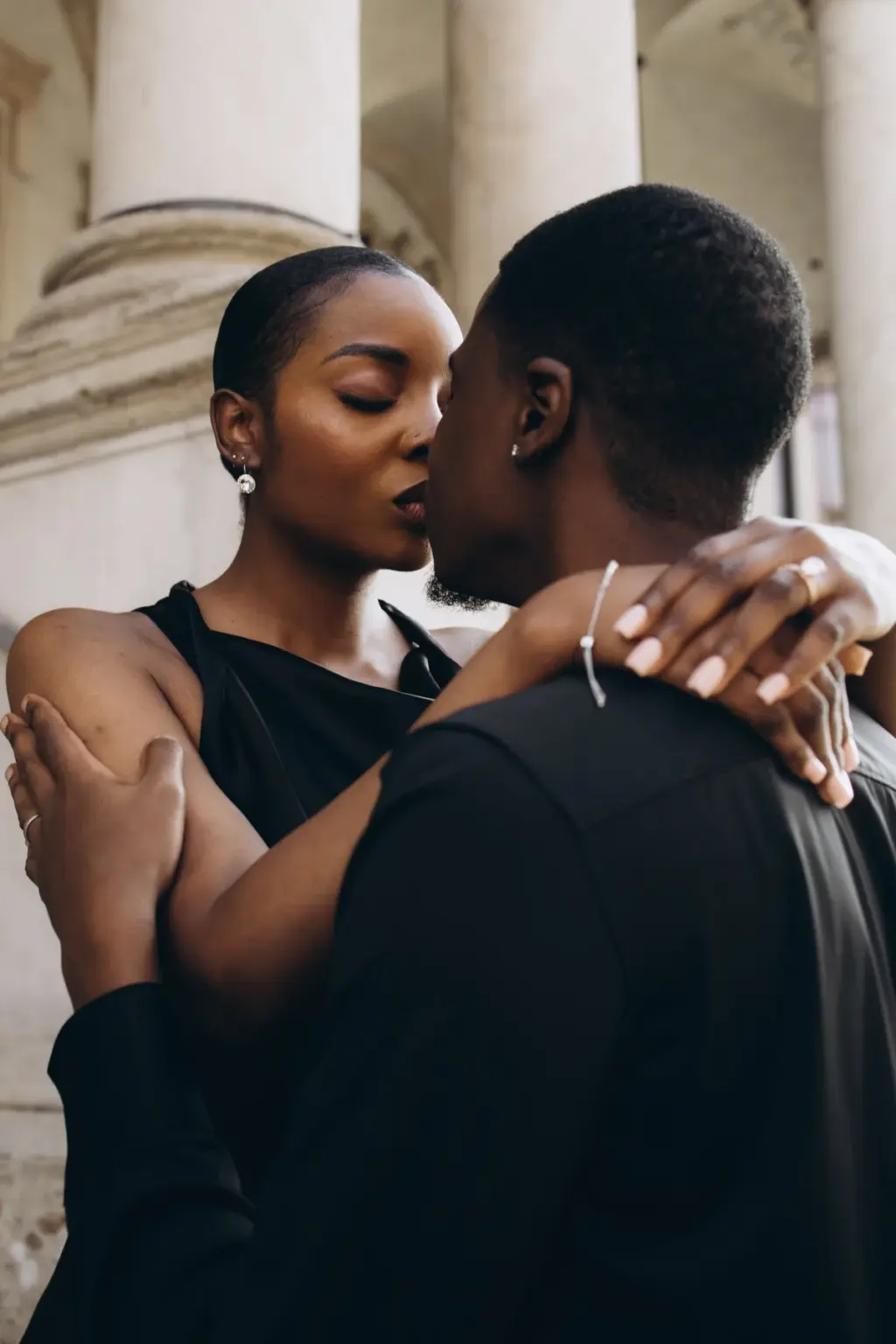 Her arms around his neck and eyes closed, she leans into him beneath tall stone columns, a thin bracelet on her wrist.