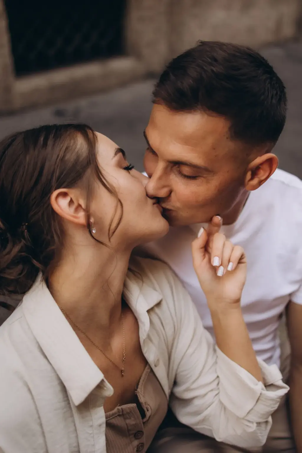 Sitting close on stone steps, a woman in a linen jacket kisses her partner, her fingertips resting lightly against his jaw.