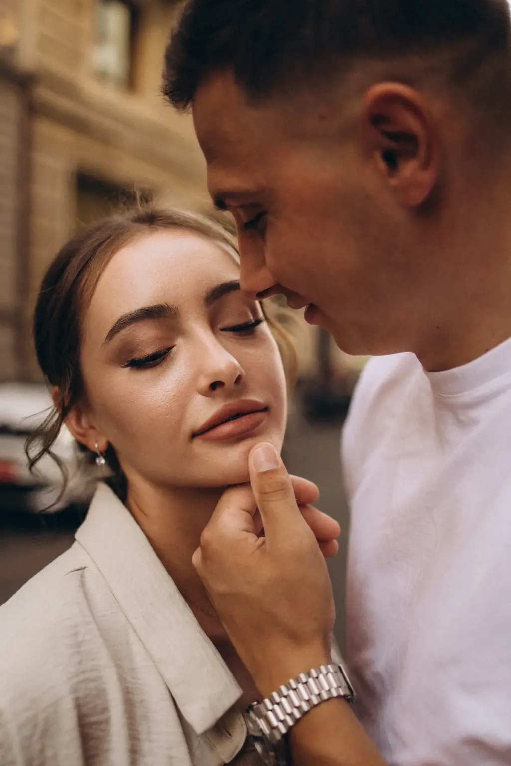 Close portrait of a woman with eyes lowered as her partner gently lifts her chin, his silver watch catching the light, on a Milan street.
