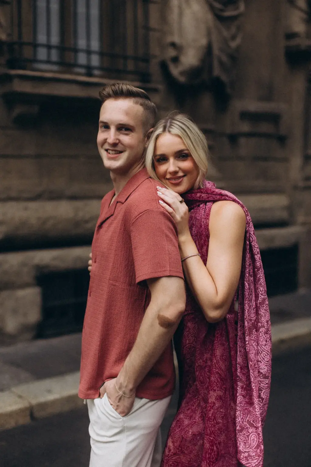 She hugs him from behind and rests her head on his shoulder as both smile at the camera beside a carved stone building.