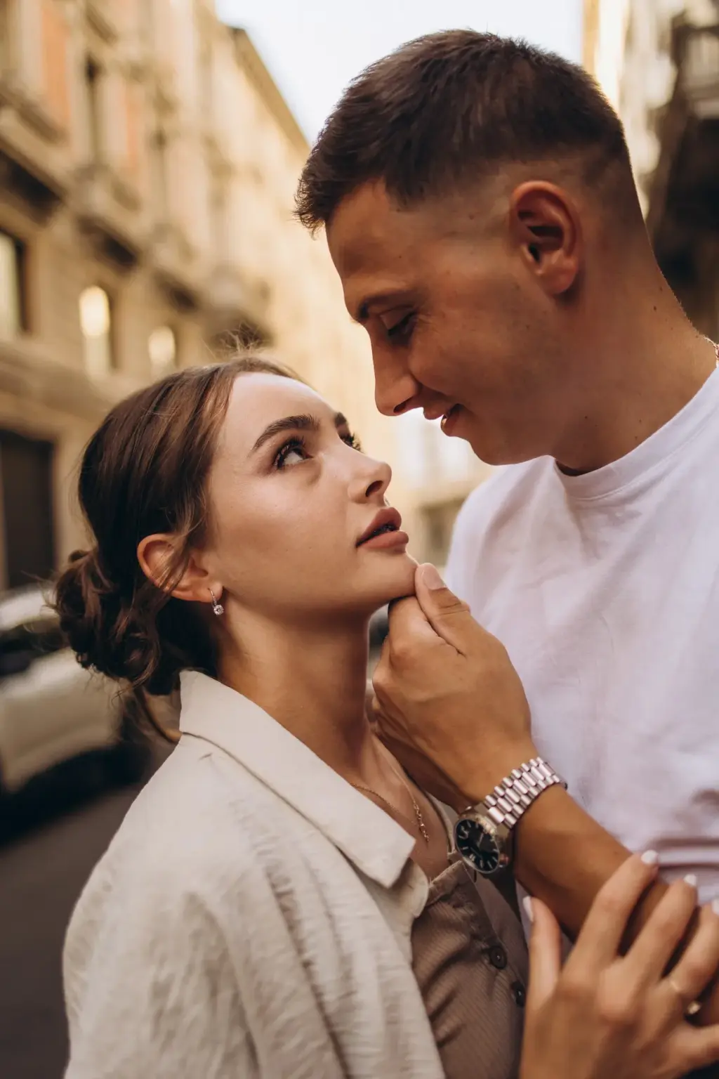 A woman with her hair in a low bun looks up at her partner as he touches her jaw and smiles, old Milan facades soft behind them.