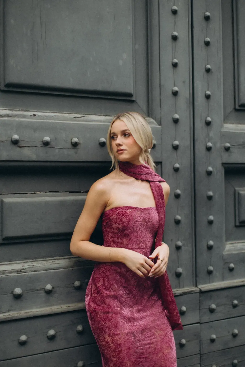 A woman in a strapless raspberry lace gown leans against a giant studded wooden door, her long scarf draped over one arm.