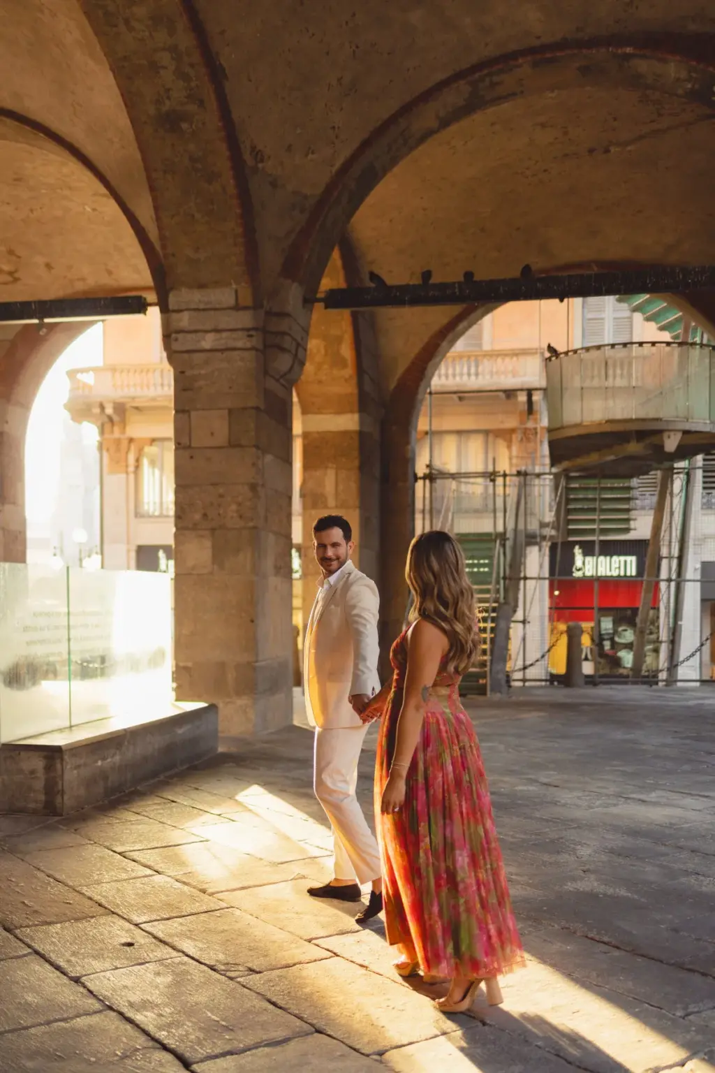 Hand in hand, they stroll through the vaulted arcade as patches of sunlight fall across the worn stone floor around them.
