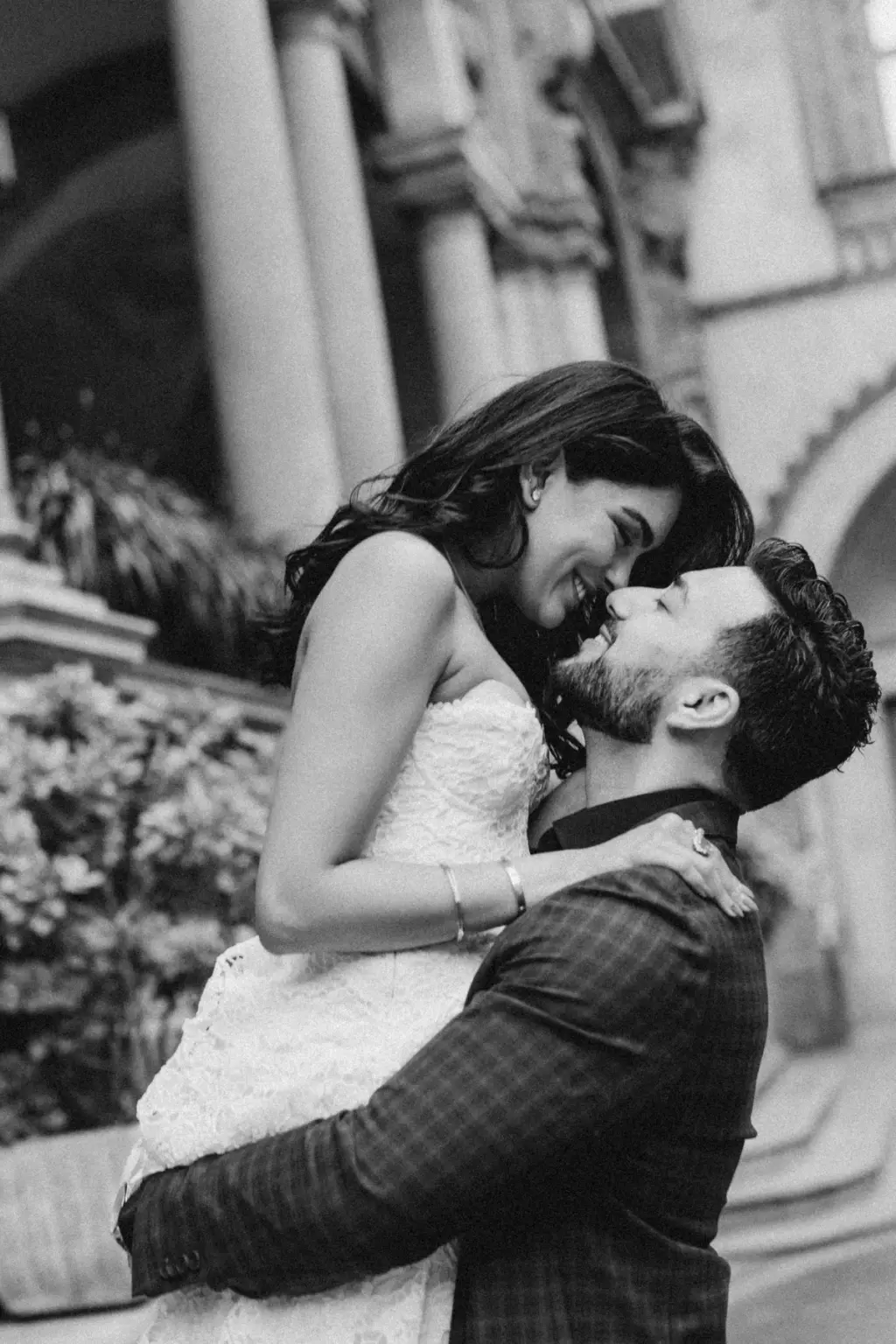 Black-and-white close view of the lift, her arms around his neck and their faces almost touching beside a carved column.