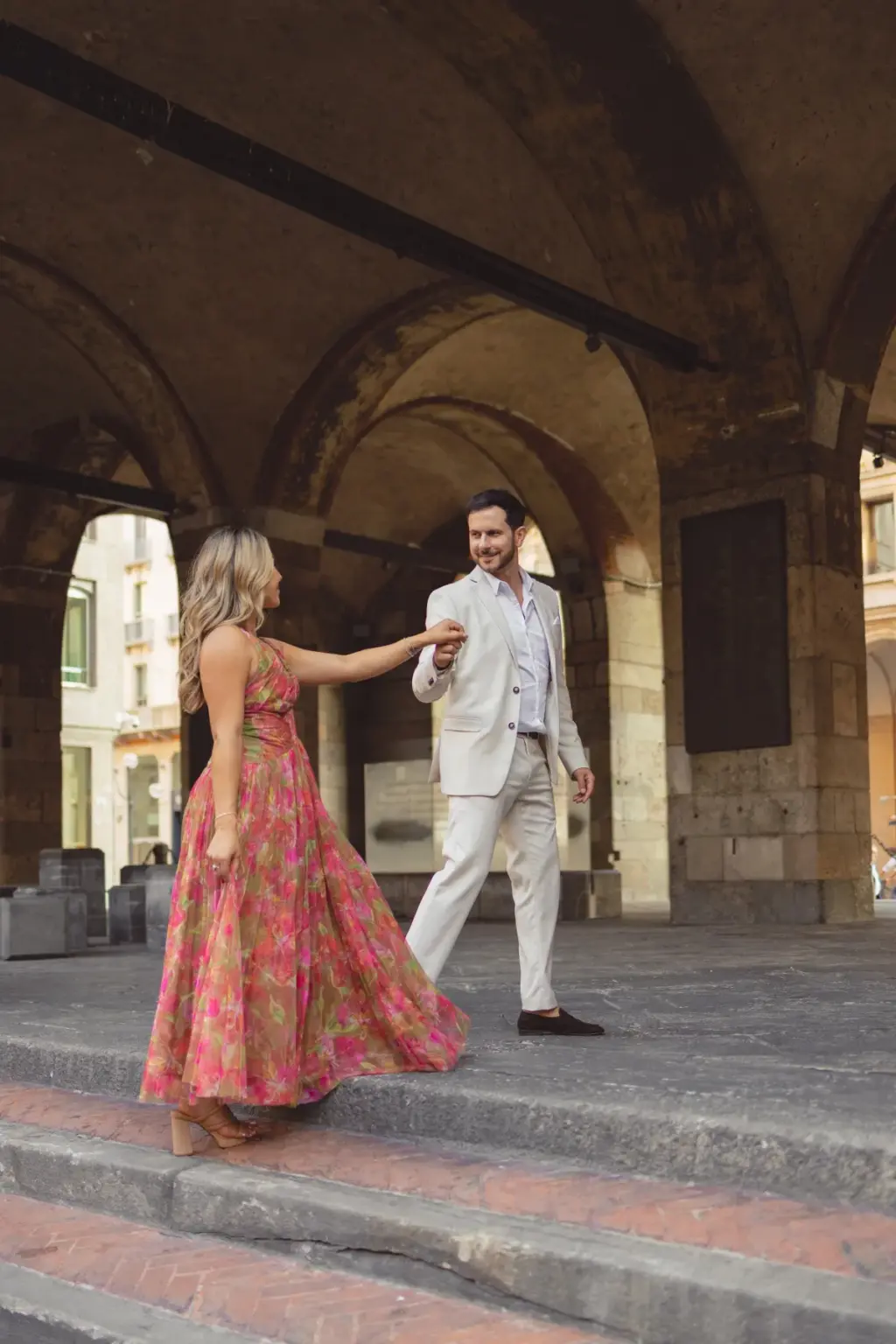 Under the stone vaults of the Piazza dei Mercanti arcade, she leads him by the hand along the low steps, dress trailing.