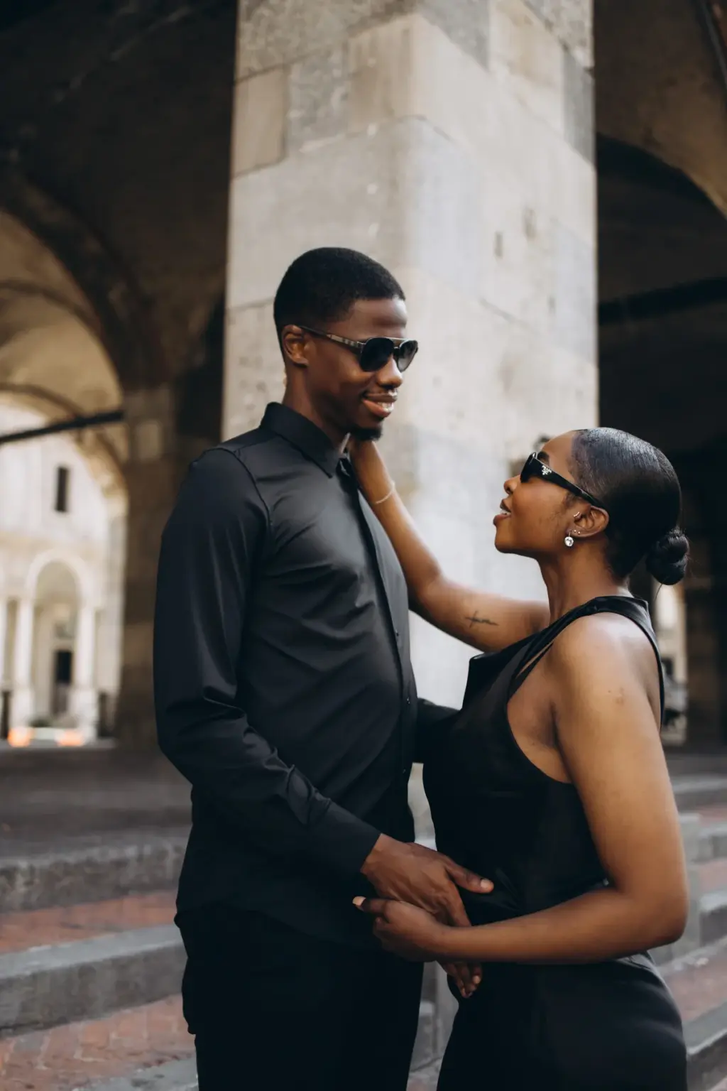 Wearing sunglasses, she reaches up to his face beneath the vaulted arcade of Piazza dei Mercanti, beside a massive stone pillar.