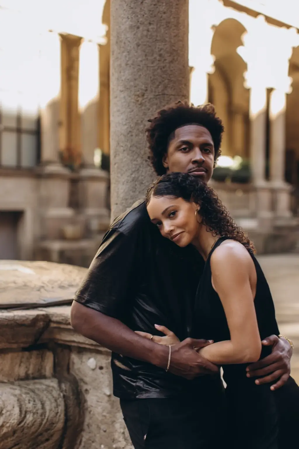 A woman rests her head on her partner's chest as he holds her close beside a stone column in the arcades of Piazza dei Mercanti.